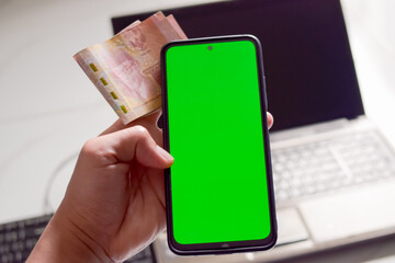 A smartphone with a green screen and Indonesian rupiah banknotes held in hand above a laptop...