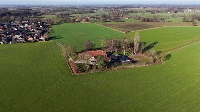 An expansive aerial drone shot of a traditional red roofed farmhouse nestled within a square of trees, surrounded by vibrant green agricultural fields and a quiet country road.