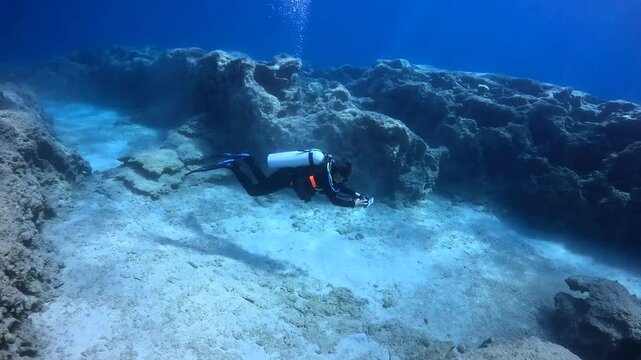 Slowly swimming scuba diver, underwater photographer. Diver in the seascape, vivid blue ocean. 
Sand and stones, clear sea and swimming underwater videographer. 
