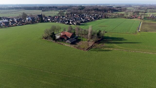 An expansive aerial drone shot of a traditional red roofed farmhouse nestled within a square of trees, surrounded by vibrant green agricultural fields and a quiet country road.