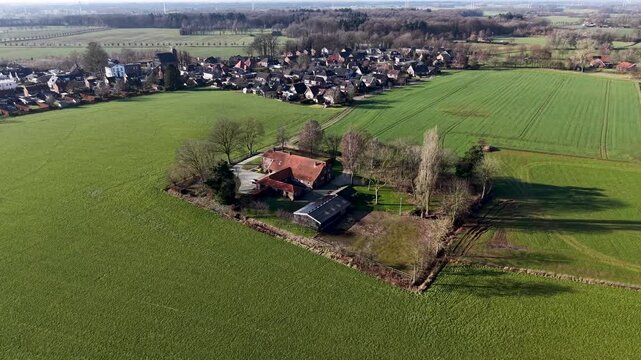 An expansive aerial drone shot of a traditional red roofed farmhouse nestled within a square of trees, surrounded by vibrant green agricultural fields and a quiet country road.
