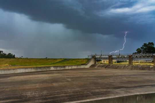 Lightning strike and heavy rain under a band of dark storm clouds over a dry reservoir spillway