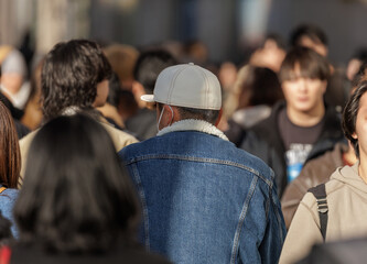 Man in denim jacket with white fleece collar and cream baseball cap walks among dappled light highlighting crowd of people moving through busy city street, evoking anonymity and city life movement. © thomasgweber