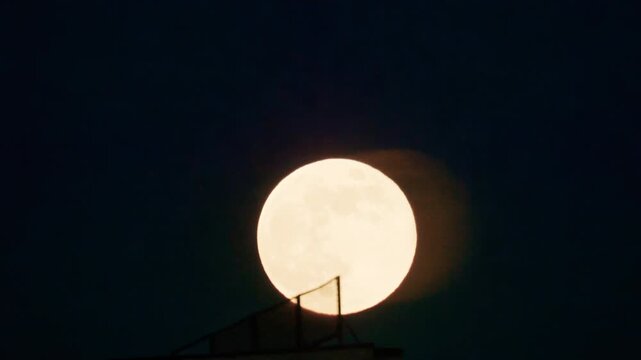 Large full moon rising in a dark night sky, partially framed by a silhouetted structure in the foreground
