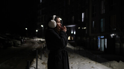 Young woman in warm earmuffs stands on night street in urban landscape. © Vasyl