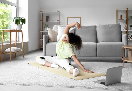 Sporty African-American woman with laptop practising pilates on yoga mat at home