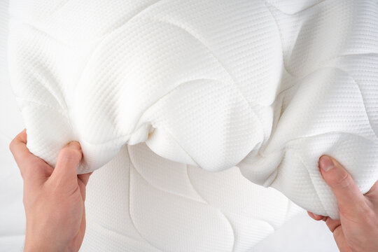 Two hands press a white quilted mattress topper, checking its soft elasticity. This highlights the comfortable, fluffy texture of the new bedding item