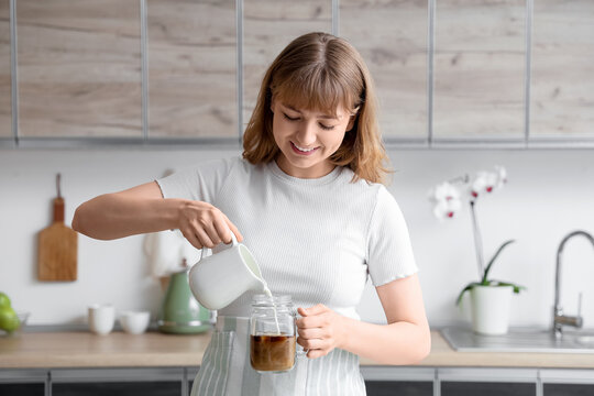 Young woman pouring milk from jug into glass with iced coffee in kitchen