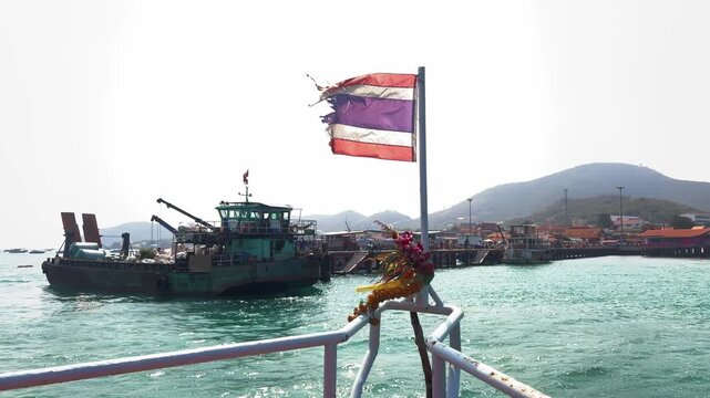 Vessel approaches busy port harbor with traditional cargo ship prominent foreground. Thai flag waves above hills bungalows dockside serene tropical setting.