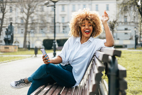 A joyful young African American woman with a blonde afro expresses excitement and laughter while sitting on a wooden bench in a public park, holding her mobile phone.
