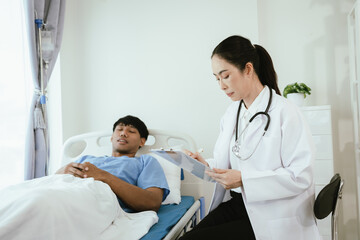 Fototapeta premium Female doctor reviewing medical records on a clipboard while a male patient rests in a hospital bed during inpatient care.