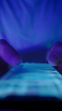 Macro view of man fingers move across a glowing touchscreen, scrolling through social media feed in the dark. Late-night smartphone use or screen dependence. Vertical video