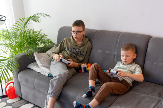 Two focused brothers in casual clothes playing video games with wireless controllers on grey sofa with colorful toys. Concept of sibling rivalry, video gaming and kids home entertainment.