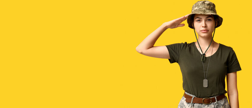 Confident young female soldier in uniform saluting on yellow background
