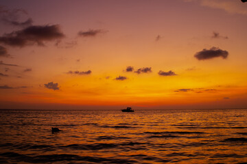 Obraz premium Golden sunset over calm sea with small boat silhouette on horizon. Warm orange sky and sea reflections creating peaceful travel landscape and serene evening atmosphere. Thailand vacation.
