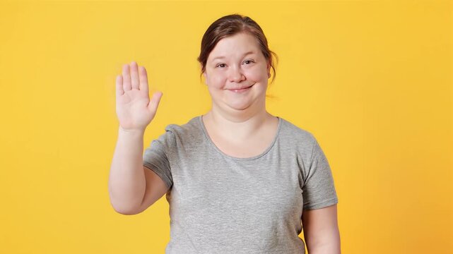 Smiling young woman with Down syndrome waving hand, looking at camera, isolated on yellow background.