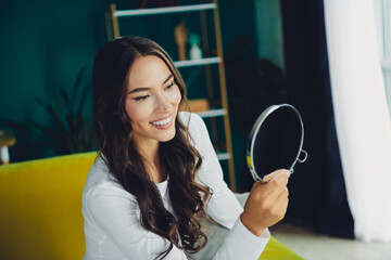 Confident young woman smiles in bright living room while checking mirror in modern cozy home...