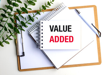 Value added phrase written on notepad with clipboard, pen and green leaves on white office table 