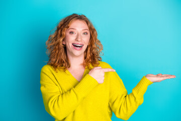 Fototapeta premium Happy redhead woman wearing a yellow sweater points to her open hand against a bright teal blue background