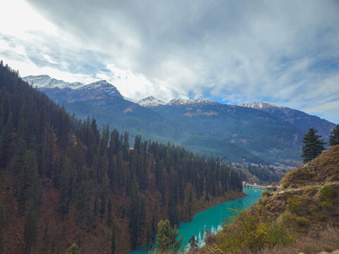 Horizontal Landscape View of Vibrant Turquoise Mountain River Winding Through Dense Pine Forests and Snowy Himalayan Peaks in Himachal Pradesh India.