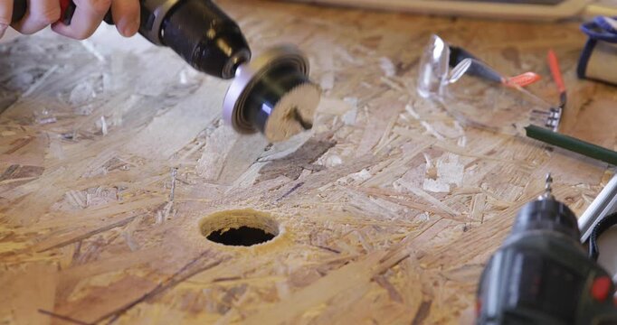 Carpenter - woodworker cuts hole through OSB plate using cordless drill and circular hole saw attachment and dusting sawdust using vacuum cleaner