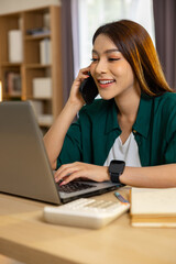 Happy freelance asian woman working with laptop at home office talking on smartphone smiling and...