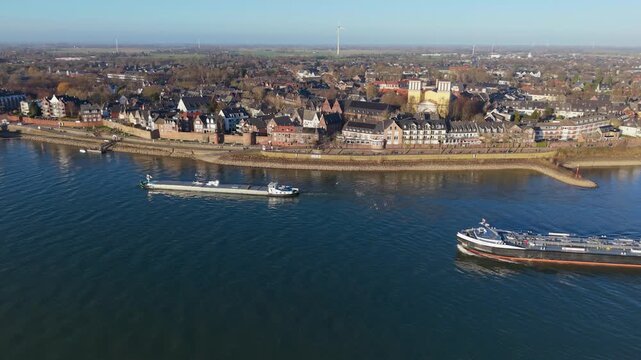 Wide aerial view of industrial cargo ships navigating a blue river alongside a historic European town. The scene features traditional architecture, riverfront promenades, and distant wind turbines.