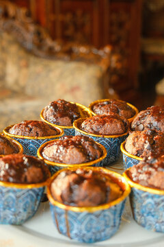 Close-up shot of freshly baked moist chocolate muffins in blue and gold patterned cups with chocolate glaze, featuring warm sunlight and a beautifully blurred vintage background.