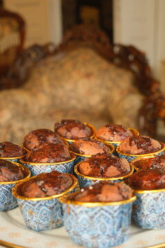 Close-up shot of freshly baked moist chocolate muffins in blue and gold patterned cups with chocolate glaze, featuring warm sunlight and a beautifully blurred vintage background.