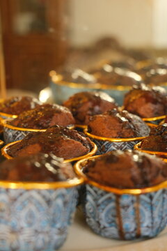 Close-up shot of freshly baked moist chocolate muffins in blue and gold patterned cups with chocolate glaze, featuring warm sunlight and a beautifully blurred vintage background.