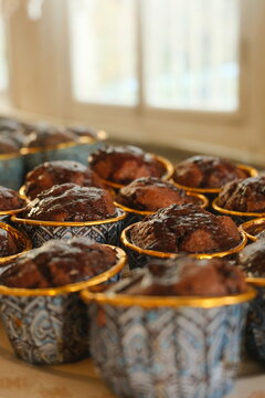 Close-up shot of freshly baked moist chocolate muffins in blue and gold patterned cups with chocolate glaze, featuring warm sunlight and a beautifully blurred vintage background.