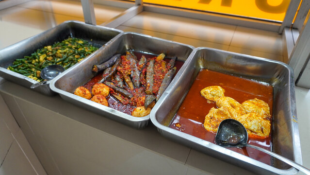 Padang food stalls share a variety of buffet menus with vegetables, chicken curry sauce, beef, eggs, tofu, tempeh. the waiter prepares the food. Menu in glass display case. Asia and asian food. Pile.
