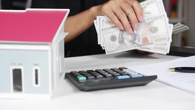 A man counts dollar bills with a miniature house in front of him. The concept of renting a house or paying taxes on an apartment. The first payment on a mortgage, rent, and home insurance.