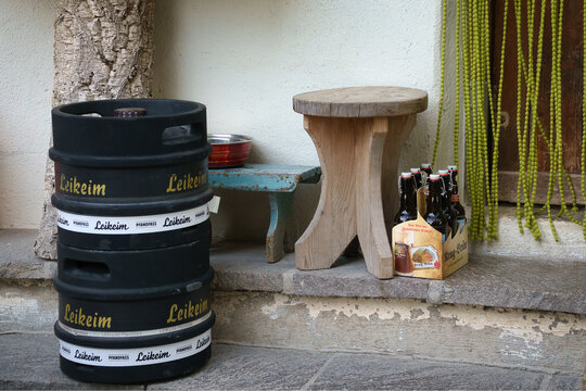 Ein kleiner Holzhocker neben einer kleinen, blauen Holzbank auf der ein metallener Trinknapf steht neben einem Sechserpack Krug-Br&auml;u Bier und einem Pfandfass mit Leikeim Bier im Vordergrund