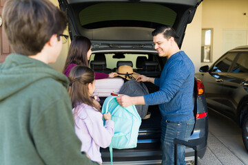 Happy family loads suitcases into car on driveway, parents and teens smiling as they prepare for a...