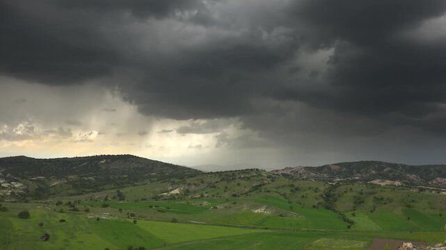 Storm clouds, rain shafts over green Balkan farmland fields, rolling wooded hills. Dark cumulonimbus, downpour curtains above lush croplands, pastures, shrub covered ridges.