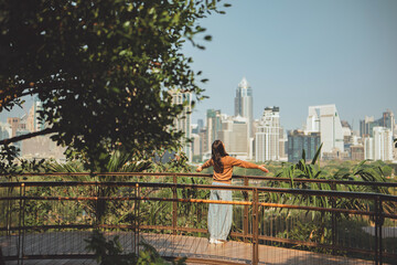 Back view of happy Asian woman standing with arms raised for freedom in green city park, Lady enjoying fresh air and wellness in urban forest, concept of mental health, stress relief, sustainable