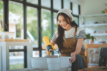 Happy asian woman using handheld dryer to dry clay pottery during ceramic workshop &ndash; smiling female enjoying creative hobby, hands-on artistic craft and peaceful leisure activity in studio environment