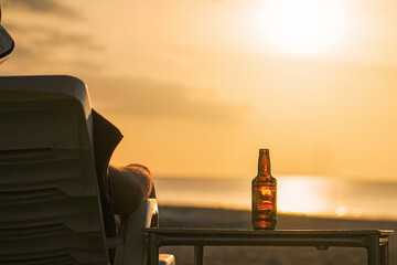 A bottle of beer on the table during sunset at sea. A man looks at the sea, rests and watches the setting sun, the concept of a holiday at a resort. Relaxing and enjoying a drink in the evening
