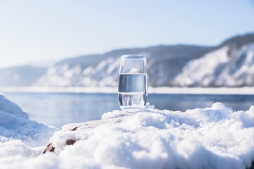 A clean glass of healthy mineral water stands in the snow in the Siberian cold. A glass filled with water stands on a snow-covered surface near a river during winter. Mountains rise in the background © Vladimir Razgulyaev