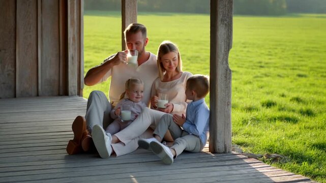 Family sitting on wooden farmhouse veranda drinking milk, morning sun, green field, relaxed weekend atmosphere. Concept for slow living and family values