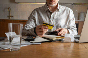 Man with credit card and wallet at his hands planning home budget at apartments kitchen with laptop...