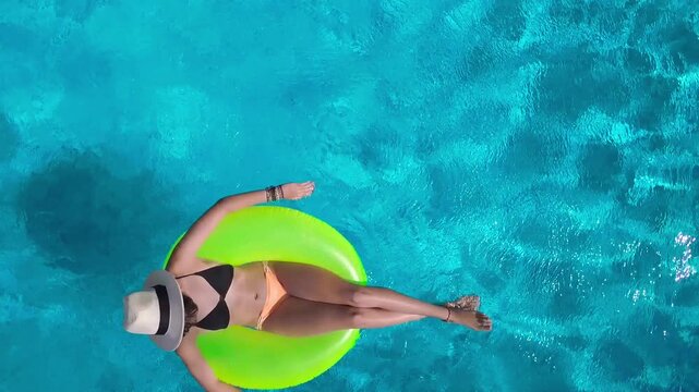 Aerial view of young woman in hat relaxing in swimming pool, beautiful girl in bikini floats on inflatable ring donut and has fun in water, tropical vacation on holiday resort, top drone view