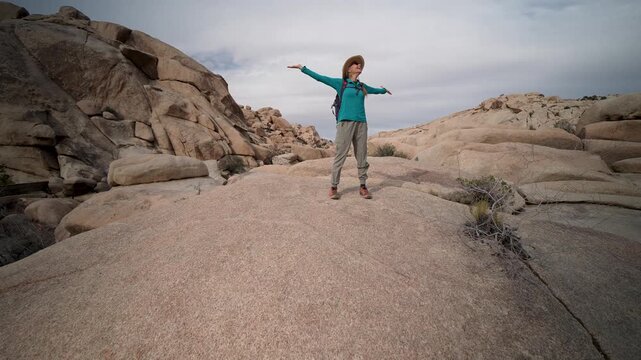 Woman hikes through Joshua Tree National Park, climbs rocks, and explores the desert landscape filled with cholla cactus and other plants.
