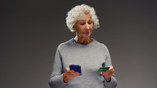Mature 70s elderly Caucasian woman senior female granny typing mobile phone hold credit banking card. Old pensioner using e-banking app for transaction. Old person holds smartphone, online shopping