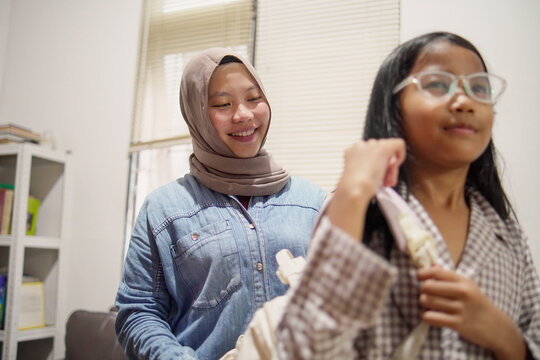 Asian Muslim mother helping her young daughter pack school supplies into her backpack, morning preparation and parenting concept