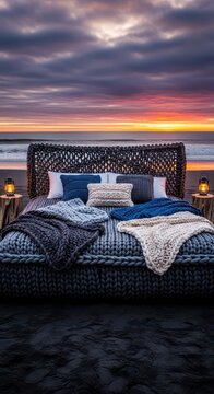 Comfortable outdoor bedding setup rests on dark sand against a dramatic ocean sunset