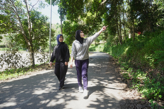 Two young Muslim women walking together for morning exercise along a green park pathway filled with trees on a bright sunny day. 