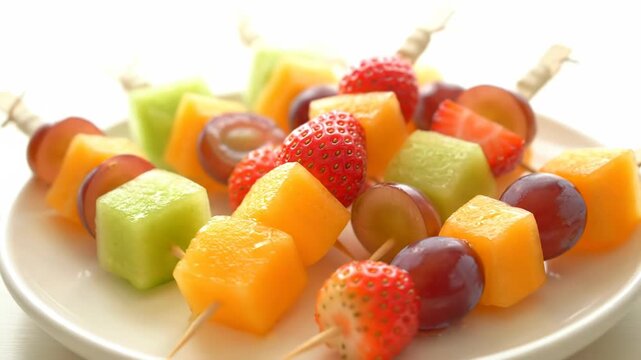 Colorful fruit skewers are arranged on a white plate. Fresh and healthy snack with melon, grapes, and strawberries.