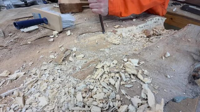 A carpenter in overalls chiselling a wooden slab in a carpentry workshop. A working man. Tools, chisel, mallet. Woodworking. Sawdust, wood chips. Real people. Lifestyle, Authentic, Candid
